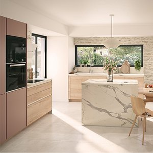 une grande cuisine claire, avec un grand îlot central. L’îlot, tout en longueur, est divisé en une moitié plan de travail en marbre, et une autre moitié étant une table en bois claire autour de laquelle sont installées 4 chaises. Des assiettes sont sur cette table pour illustrer que l’on peut s’y attabler pour un repas.