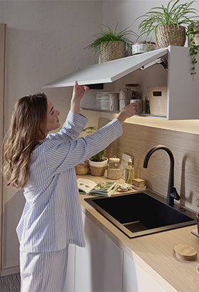 Femme en pyjama rayé ouvre un placard en hauteur dans une cuisine contemporaine avec plantes vertes, évier noir et plan de travail clair