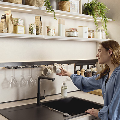 Cuisine contemporaine avec étagères remplies de bocaux, tablette fixée au mur et femme plaçant des tasses au-dessus d’un évier noir