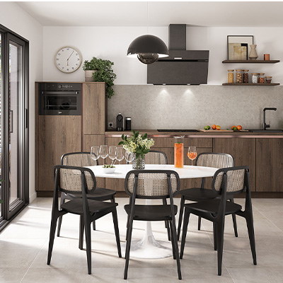 une cuisine de style industriel, aux murs blancs avec placards en bois foncé, aux accents en métal noir. Au premier plan, une table et des chaises noires avec empiècement en rotin clair. La décoration est minimaliste, avec quelques plantes vertes et des étagères ouvertes sur lesquelles figurent des bocaux en verre.